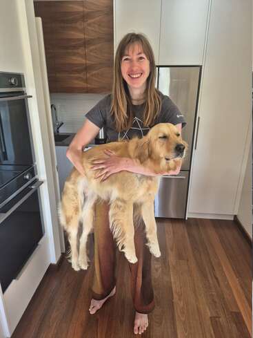 Une femme souriante, pieds nus, se tient debout dans une cuisine moderne, tenant joyeusement dans ses bras un golden retriever qui semble détendu. Le chien a l'air satisfait et somnolent.