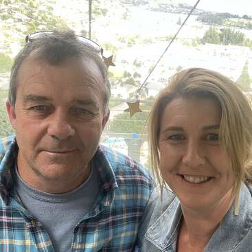 A imagem mostra um homem e uma mulher posando para uma selfie, com uma vista panorâmica de árvores e edifícios ao fundo, provavelmente tirada ao ar livre durante o dia.