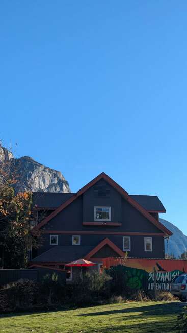 Uma casa escura com acabamento vermelho está sob um céu azul claro. As montanhas arborizadas se erguem atrás de você. Um guarda-chuva vermelho e um mural colorido acrescentam vibração à cena.