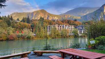 Uma cena serena à beira do rio apresenta mesas de piquenique de madeira em primeiro plano, uma floresta exuberante, árvores coloridas de outono, um rio tranquilo, edifícios distantes e majestosas montanhas iluminadas pelo sol.