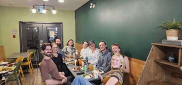 Um grupo de nove amigos se senta à mesa de um restaurante, sorrindo e desfrutando de uma refeição juntos. O ambiente é aconchegante, com paredes verdes e iluminação quente.