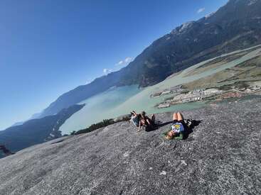 Três pessoas relaxam em um parapeito de uma montanha rochosa, com vista para um vale panorâmico com um rio, edifícios industriais e montanhas verdes imponentes sob um céu azul claro.