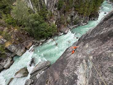 Um alpinista de laranja escala uma face íngreme de rocha acima de um rio azul-turquesa, cercado por uma densa floresta verde e penhascos rochosos escarpados, mostrando aventura.