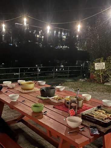 Uma mesa de piquenique vermelha está preparada para uma refeição ao ar livre à noite. Luzes de cordas brilham acima e edifícios refletem na água ao fundo. Um ambiente aconchegante.