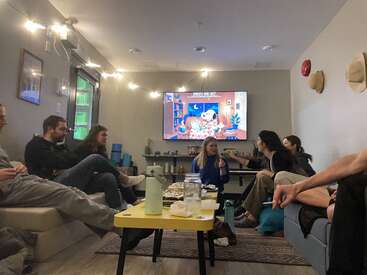 Um grupo de amigos se senta confortavelmente em uma sala de estar aconchegante, conversando e comendo, enquanto um desenho animado chamado "Snoopy e Jazz" passa na TV.