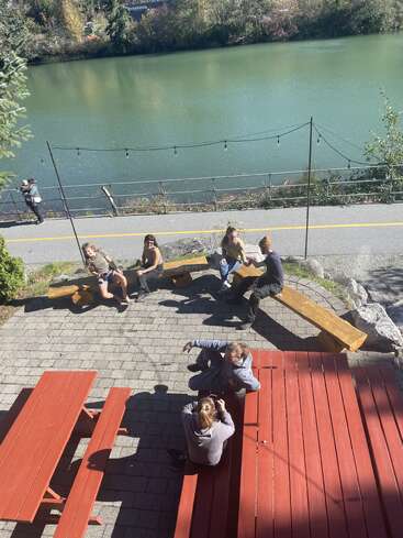 Várias pessoas sentadas em bancos e mesas de piquenique vermelhas à beira de um rio, aproveitando o clima ensolarado. As árvores e a água criam uma atmosfera tranquila ao ar livre, com luzes de cordas no alto.