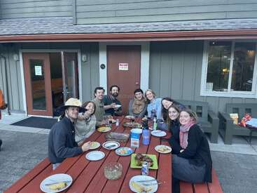 Um grupo de nove amigos senta-se ao redor de uma mesa de piquenique vermelha ao ar livre, compartilhando uma refeição e sorrindo. Alimentos e bebidas estão espalhados pela mesa. Ambiente aconchegante.
