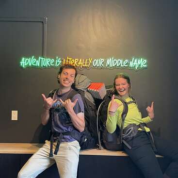 Dois jovens adultos sorridentes com mochilas posam sob uma placa de neon onde se lê "Adventure is literally our middle name" (Aventura é literalmente nosso nome do meio), fazendo sinais com a mão em forma de shaka e parecendo animados para a aventura.