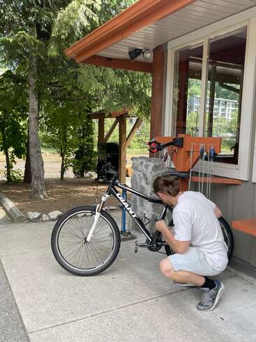 Uma pessoa vestindo camisa clara e bermuda está consertando uma bicicleta do lado de fora, próximo a um prédio cercado por árvores, usando ferramentas de uma estação de trabalho laranja.
