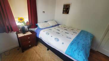 Um quarto aconchegante apresenta uma cama bem arrumada com roupas de cama azuis e brancas, uma mesa de cabeceira de madeira com um vaso de girassol, cortinas vermelhas, janela clara e arte na parede.
