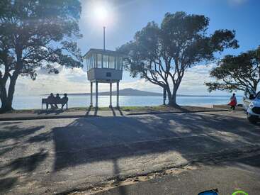 L'image montre une scène de bord de mer ensoleillée avec une tour de sauvetage, des personnes assises sur un banc, des arbres, une camionnette et de l'eau avec une île au loin.