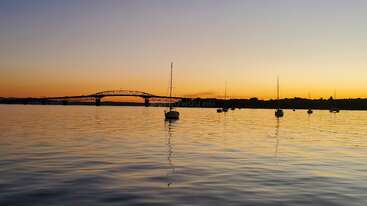 Cette image sereine capture un coucher de soleil paisible sur des eaux calmes, avec des voiliers flottant doucement. Un pont lointain enjambe l'horizon, se découpant sur le ciel jaune orangé.