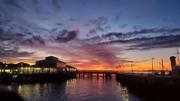 Un coucher de soleil vibrant illumine le ciel de teintes orange et violettes au-dessus d'une jetée tranquille, reflétant les lumières sur l'eau calme, créant une atmosphère sereine.