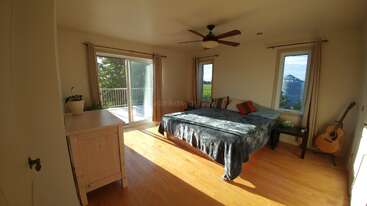 Chambre lumineuse avec parquet, grand lit, ventilateur de plafond, portes coulissantes donnant sur un balcon, commode, plante en pot et guitare acoustique.