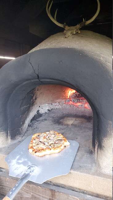 A rustic wood-fired oven with a crack above the entrance bakes a homemade pizza. Flames glow inside. Deer antlers decorate the oven’s top, creating a cozy atmosphere.