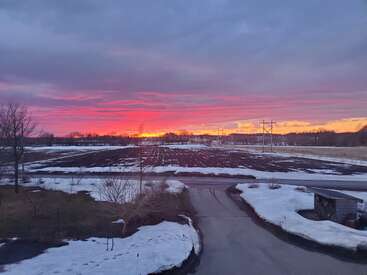 A stunning sunset casts vivid pink, orange, and purple hues across the sky above a rural snowy landscape, barren field, trees, and a paved road.