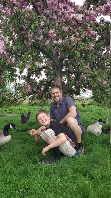 A man and a boy kneel on lush grass beneath a blooming pink tree, surrounded by geese and a chicken, sharing a joyful outdoor moment together.