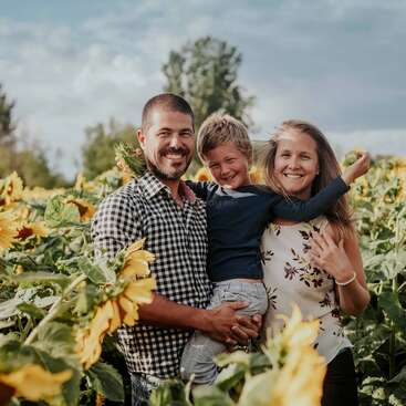 Eine lächelnde Familie steht zusammen in einem leuchtenden Sonnenblumenfeld unter einem teilweise bewölkten Himmel. Der Vater hält sein Kind im Arm, alle strahlen Glück und Wärme aus.