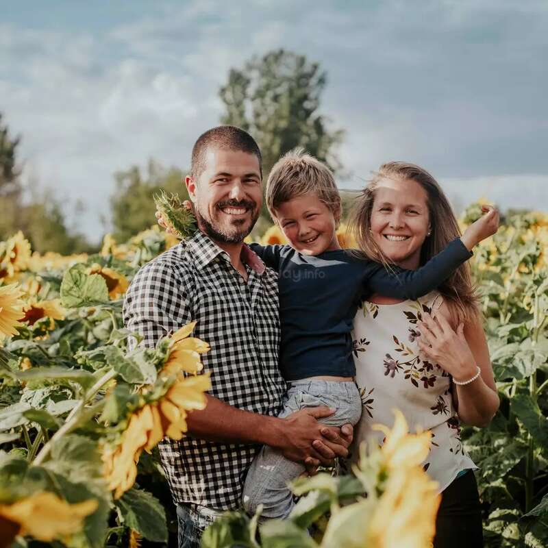 A smiling family stands together in a vibrant sunflower field under a partly cloudy sky. The father holds their child, all radiating happiness and warmth.