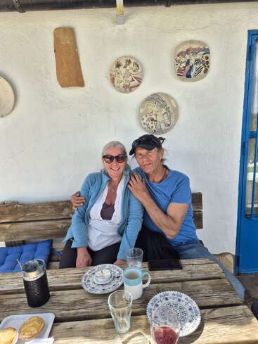 Una pareja sonriente se sienta muy junta en un banco rústico de madera, disfrutando de un día soleado, bebidas y pasteles. Detrás de ellos, cuelgan platos decorativos de la pared blanca.