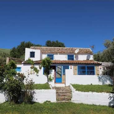 This image shows a charming white house with blue windows and doors, surrounded by greenery, under a clear blue sky, evoking a peaceful Mediterranean countryside ambiance.