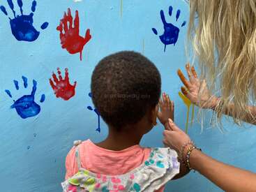 Un niño y un adulto crean juntos huellas de manos de colores en una pared azul. El adulto guía la mano del niño, fomentando la creatividad, el aprendizaje y la colaboración alegre a través del arte.