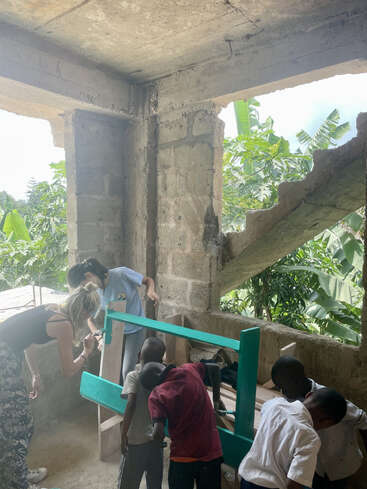 En un edificio parcialmente construido, dos mujeres y tres niños pintan juntos una estructura de madera verde. La luz del sol entra a raudales; en el exterior se ve una exuberante vegetación.