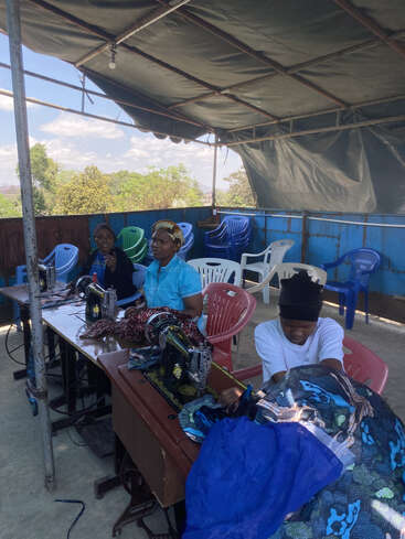 Tres mujeres están sentadas ante máquinas de coser antiguas, trabajando con telas de colores bajo un toldo improvisado. Al fondo, sillas de plástico apiladas y árboles verdes.