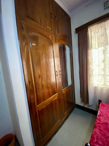 Esta imagen muestra un armario de madera con varias puertas en un rincón de un dormitorio. Se ve una ventana con cortinas y parte de una cama.