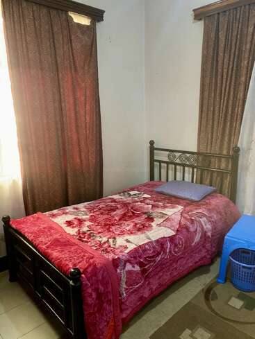 Un dormitorio sencillo con una cama individual con una manta de flores rojas, una almohada azul, dos cortinas estampadas, una mesa de plástico azul y un cesto tejido para la ropa sucia.