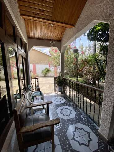 Una acogedora veranda con baldosas estampadas, techo de madera, sillas, barandilla, una maceta y exuberante vegetación exterior. La luz natural ilumina la tranquila zona de estar al aire libre.
