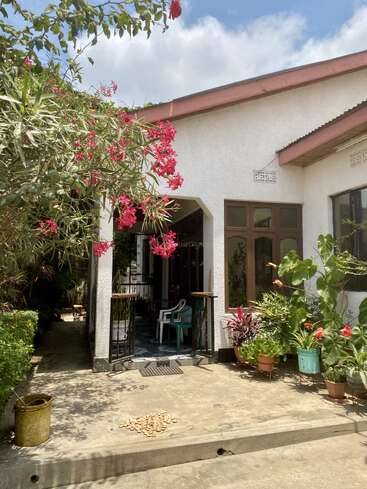 Una pintoresca casa blanca con jardín, vibrantes flores rosas, plantas en macetas y sillas de plástico en el porche bajo un cielo azul parcialmente nublado.