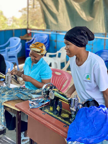 Two women are seated at sewing machines, working with colorful fabrics. One woman wears a headscarf, the other a black hat. Plastic chairs fill the background.