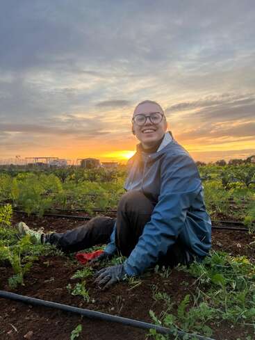 Uma pessoa usando óculos e uma jaqueta azul senta-se alegremente em uma horta ao nascer do sol, cercada por plantas e terra, aproveitando a manhã tranquila.