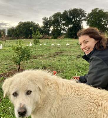 Eine lächelnde Frau sitzt draußen auf einer Wiese, im Vordergrund ist ein großer, flauschiger weißer Hund zu sehen. Im Hintergrund schwimmen Enten in der Nähe kleiner Bäume umher.