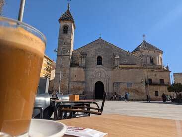 Auf einem Café-Tisch auf einem sonnigen Platz steht eine Tasse Kaffee, im Hintergrund sieht man eine alte Steinkirche und ein paar Leute. Es herrscht eine ruhige Atmosphäre.