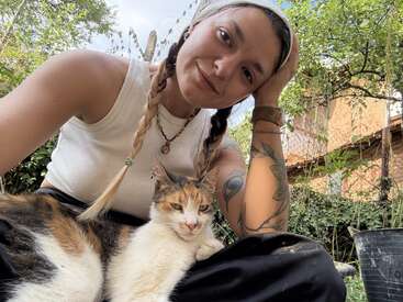 Eine Frau mit zwei Zöpfen und Tattoos sitzt draußen, lächelt sanft, trägt ein weißes Tanktop und hält eine entspannte Dreifarbkatze auf dem Schoß. Im Hintergrund ein üppiger Garten.