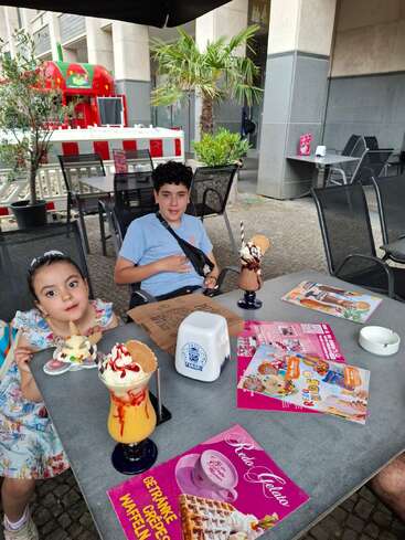 Dos niños sentados en la mesa de una cafetería al aire libre disfrutan de coloridos helados y bebidas. Los menús están desplegados, y hay plantas de palmera y un camión rojo de comida cerca.