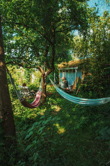 The image depicts a serene backyard scene with two hammocks suspended between trees, a person relaxing in one, and a small shed in the background, surrounded by lush greenery.