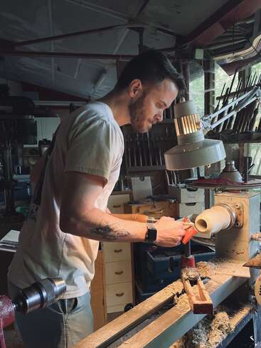 The image depicts a man intently working on a piece of wood using a lathe in a cluttered workshop, surrounded by various tools and equipment.