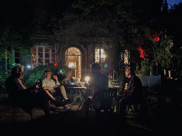 The image depicts a group of people gathered around a table in a garden at night, enjoying drinks and conversation. The atmosphere is relaxed and social.