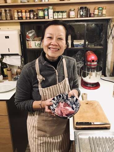 The image depicts a woman in a kitchen, wearing an apron and holding a bowl of raw meat, with a cutting board and knife nearby, in a well-organized space.