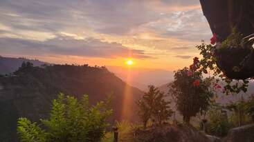 A stunning sunrise illuminates lush green mountains, vibrant flowers, and hanging plants. Soft orange and pink clouds create a peaceful, serene landscape perfect for reflection.