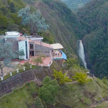 A beautiful mountainside building overlooks lush green valleys, with a scenic waterfall in the distance. The tranquil setting blends architecture with stunning natural beauty.