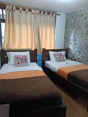 This cozy room features two single beds with colorful, embroidered pillows, brown and orange bedding, light curtains, wooden floors, and a rustic stone accent wall.