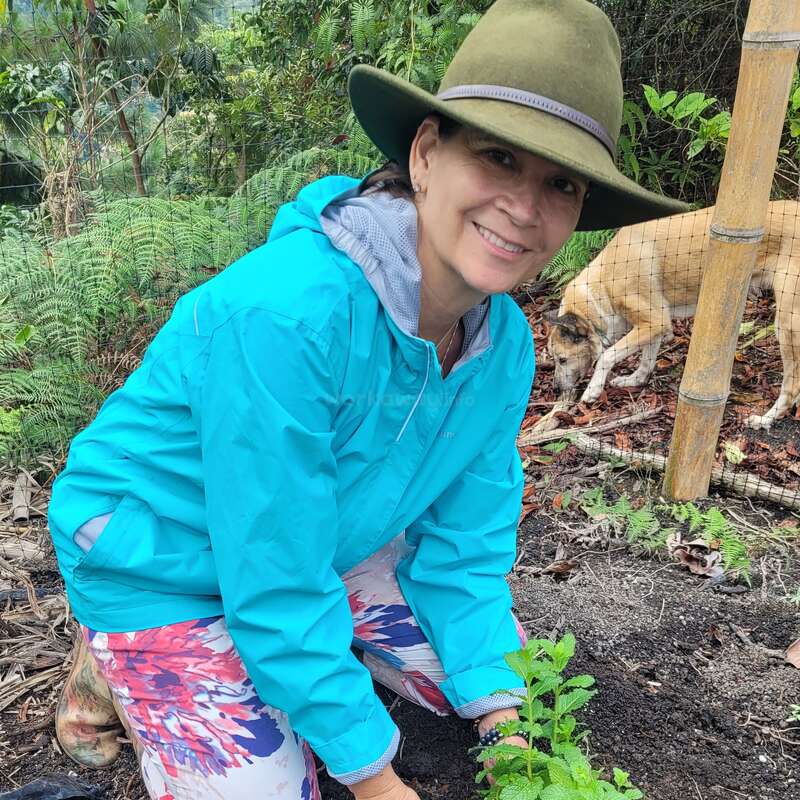 A smiling woman wearing a green hat and blue jacket kneels in a garden, planting a young green plant. A dog sniffs the ground nearby.