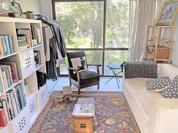 A cozy sunlit room features a cream sofa, armchair, bookshelf, garment rack, floral rug, and large window overlooking a green, wooded outdoor view. Relaxing atmosphere.