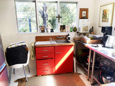A cozy kitchen with red cabinets, sunlight streaming through large windows, various utensils on a metal countertop, plants outside, and eclectic decorations and framed artwork.