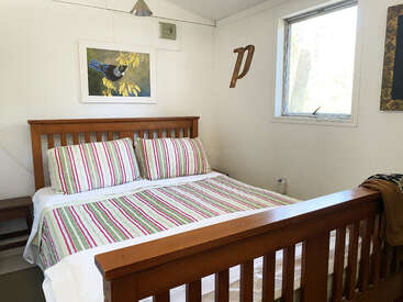 This cozy bedroom features a wooden bed with striped bedding, white walls, a window, bird artwork, a wooden “P” decoration, and a side table.