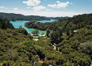 The image depicts a serene landscape featuring a body of water, lush greenery, and a few buildings, set against a backdrop of rolling hills and a blue sky with clouds.